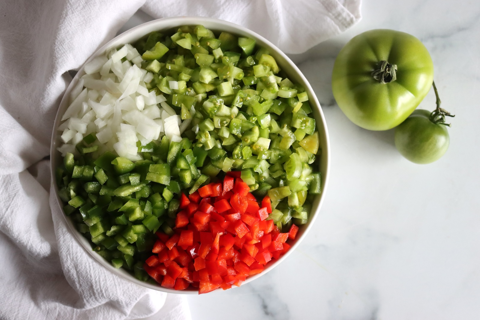 Bowl of diced green tomatoes, green peppers, red peppers, and onion beside whole green tomatoes