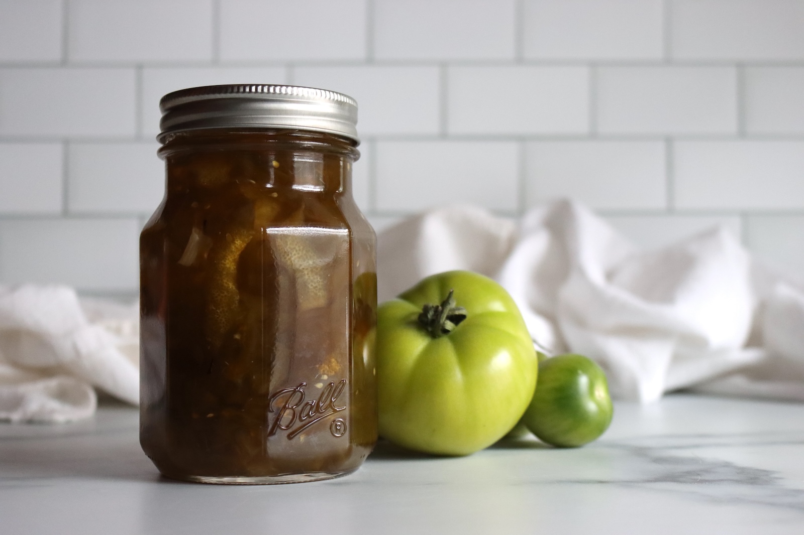 Pint jar of Italian green tomato preserves beside fresh green tomatoes on a marble counter