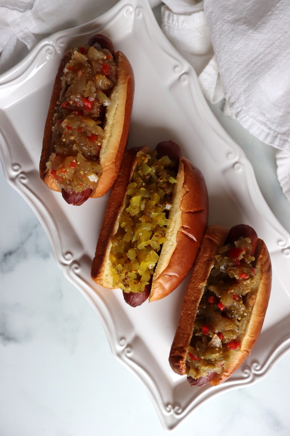 Three hot dogs on a platter topped with classic, dilled, and hot dog green tomato relish