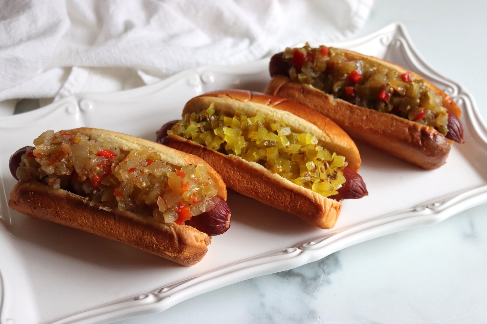 Three hot dogs topped with three different green tomato relishes on a white serving platter