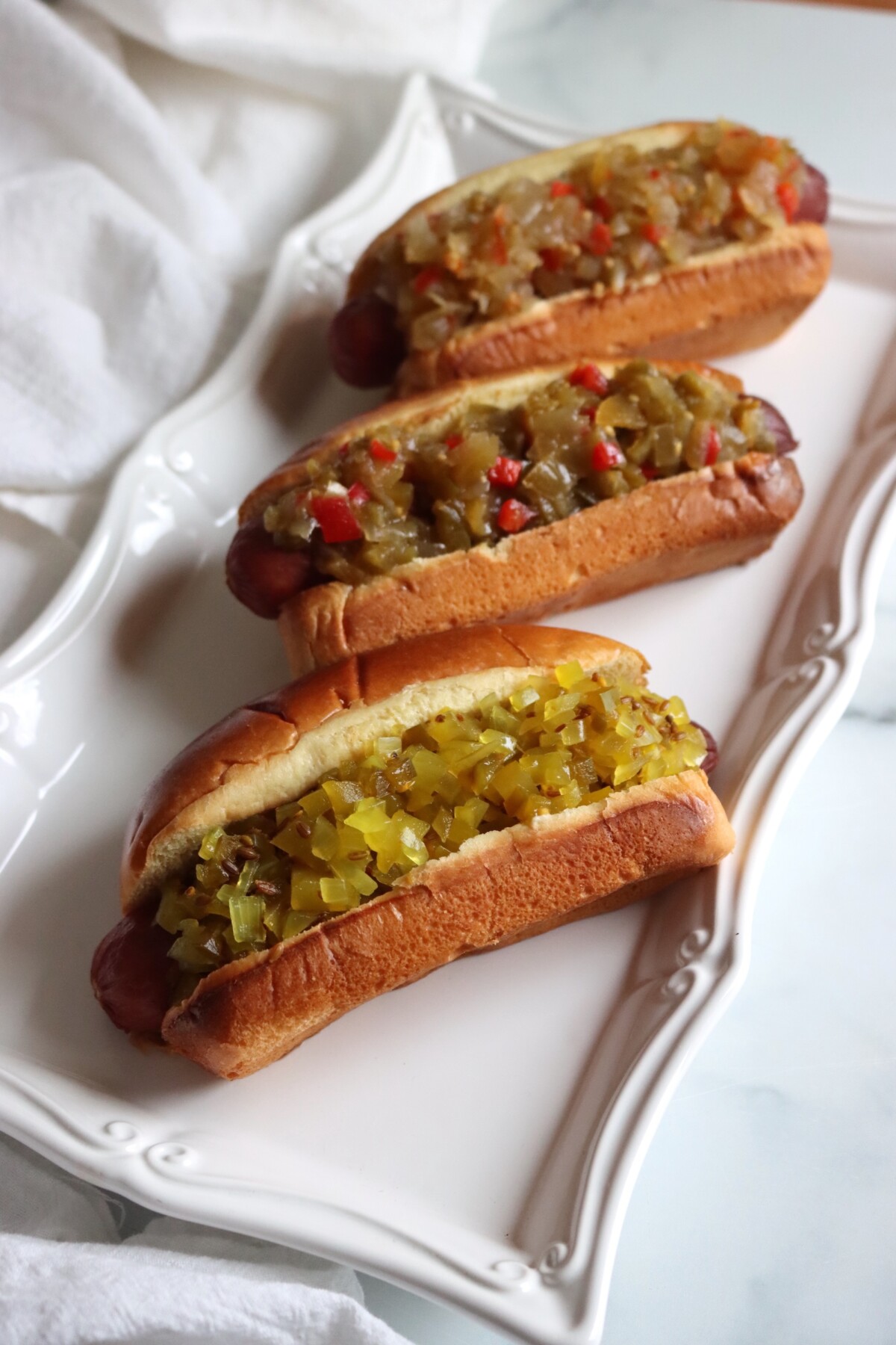 Platter of three hot dogs with green tomato relishes, the front one topped with dilled green tomato relish