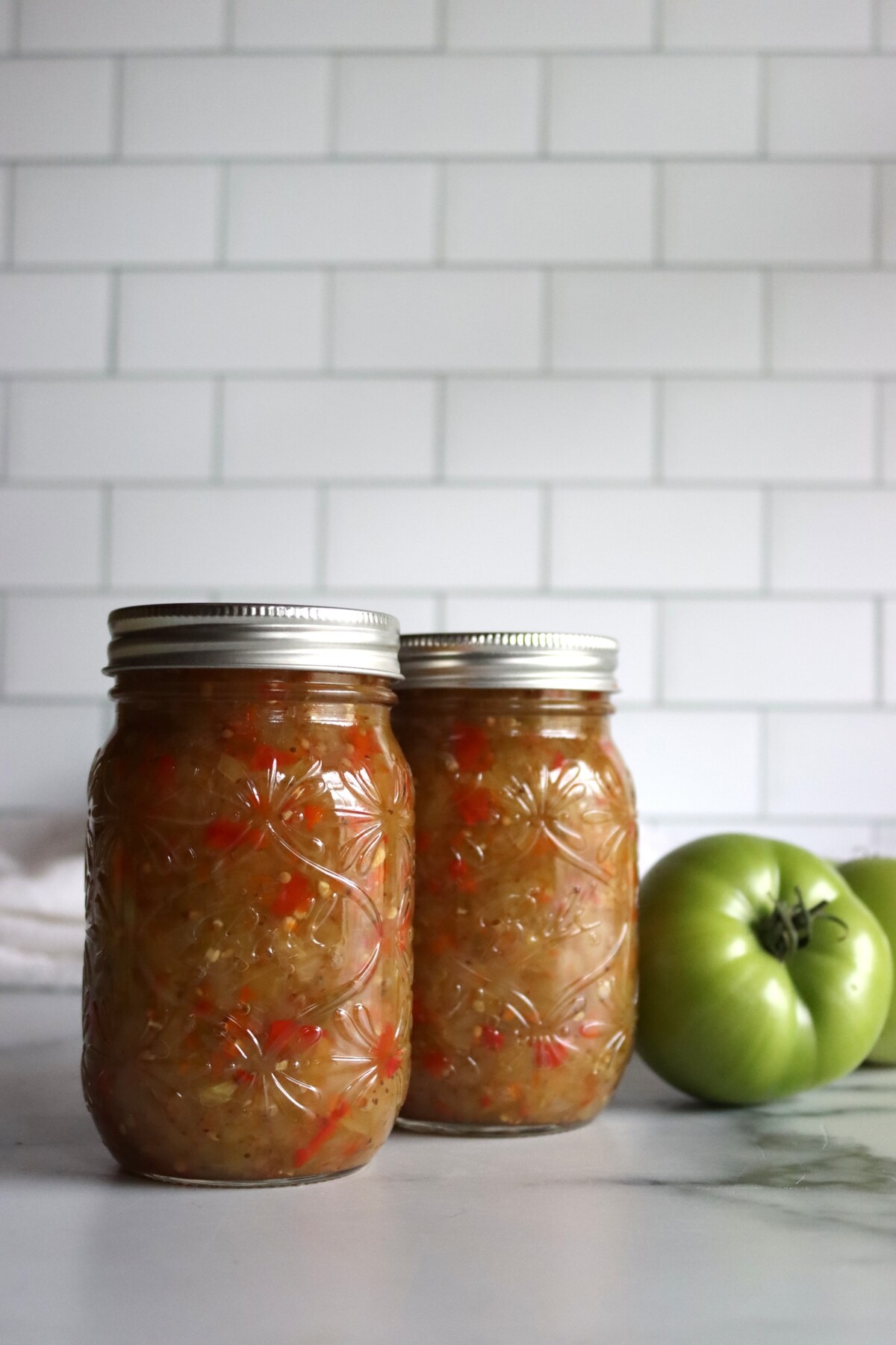 Two jars of green tomato relish with red pepper flecks next to a whole green tomato