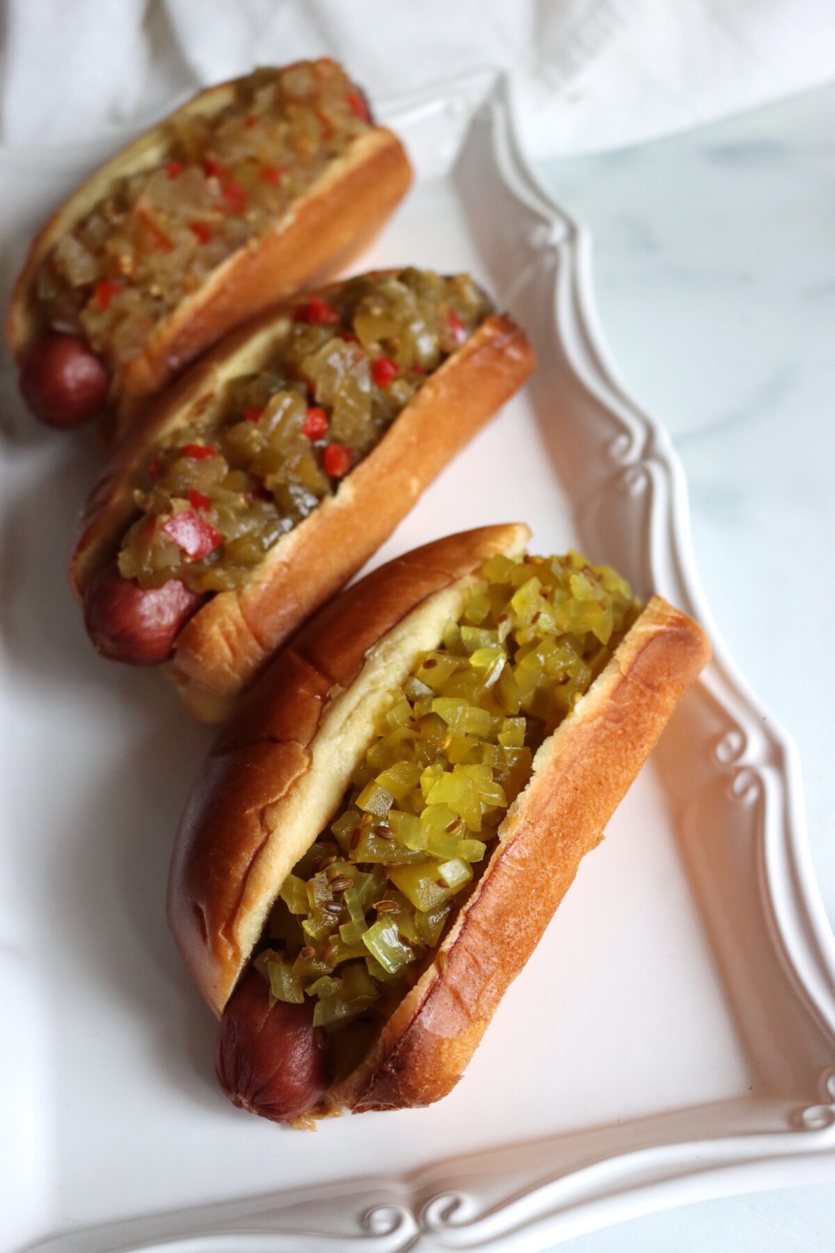 Three hot dogs on a white platter topped with green tomato relishes, with dilled green tomato relish in front