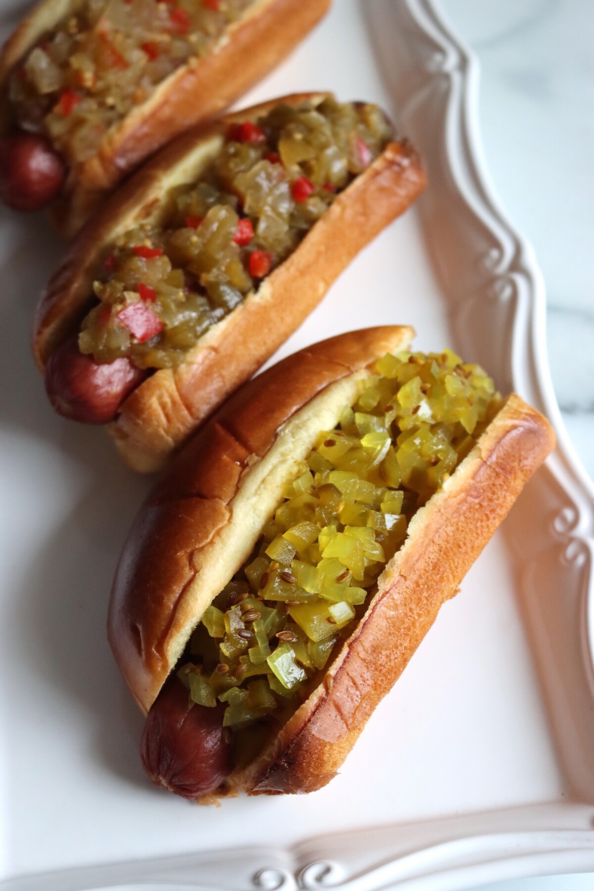 White platter of hot dogs topped with green tomato relishes, with dilled green tomato relish on the front hot dog