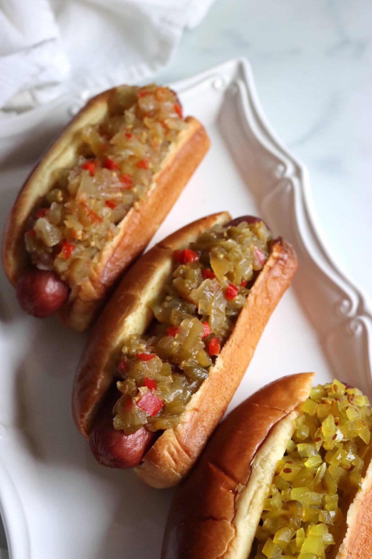Three hot dogs on a white platter topped with green tomato relishes, with hot dog relish in the center