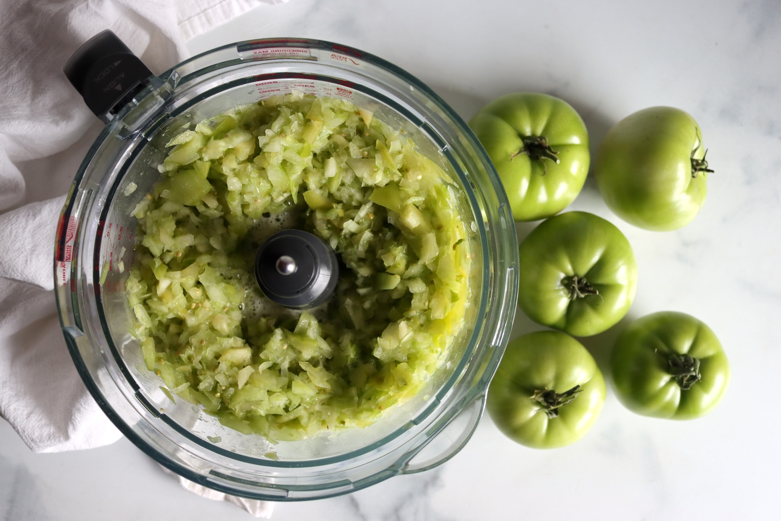Finely chopped green tomatoes in a food processor bowl beside whole green tomatoes