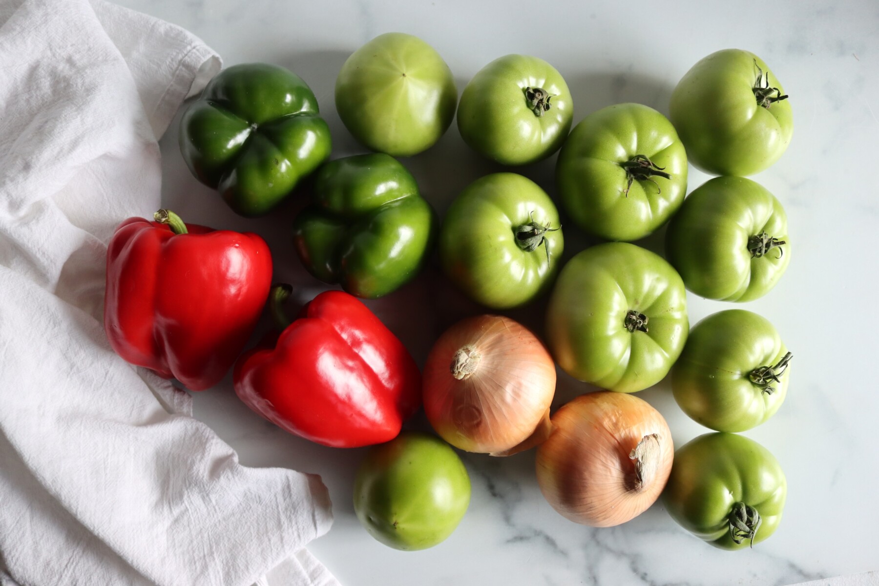 Green tomatoes, red and green bell peppers, and onions on a marble counter for making relish