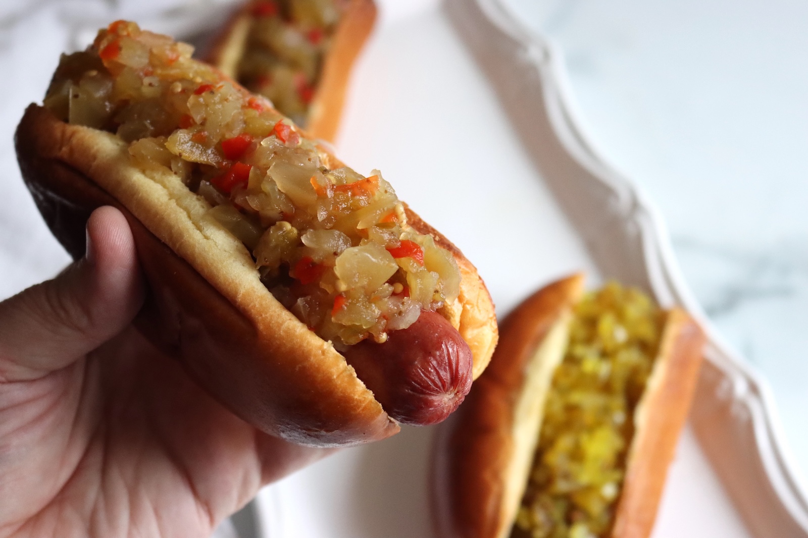 Hand holding a hot dog topped with green tomato relish, with more relish-topped hot dogs on a platter