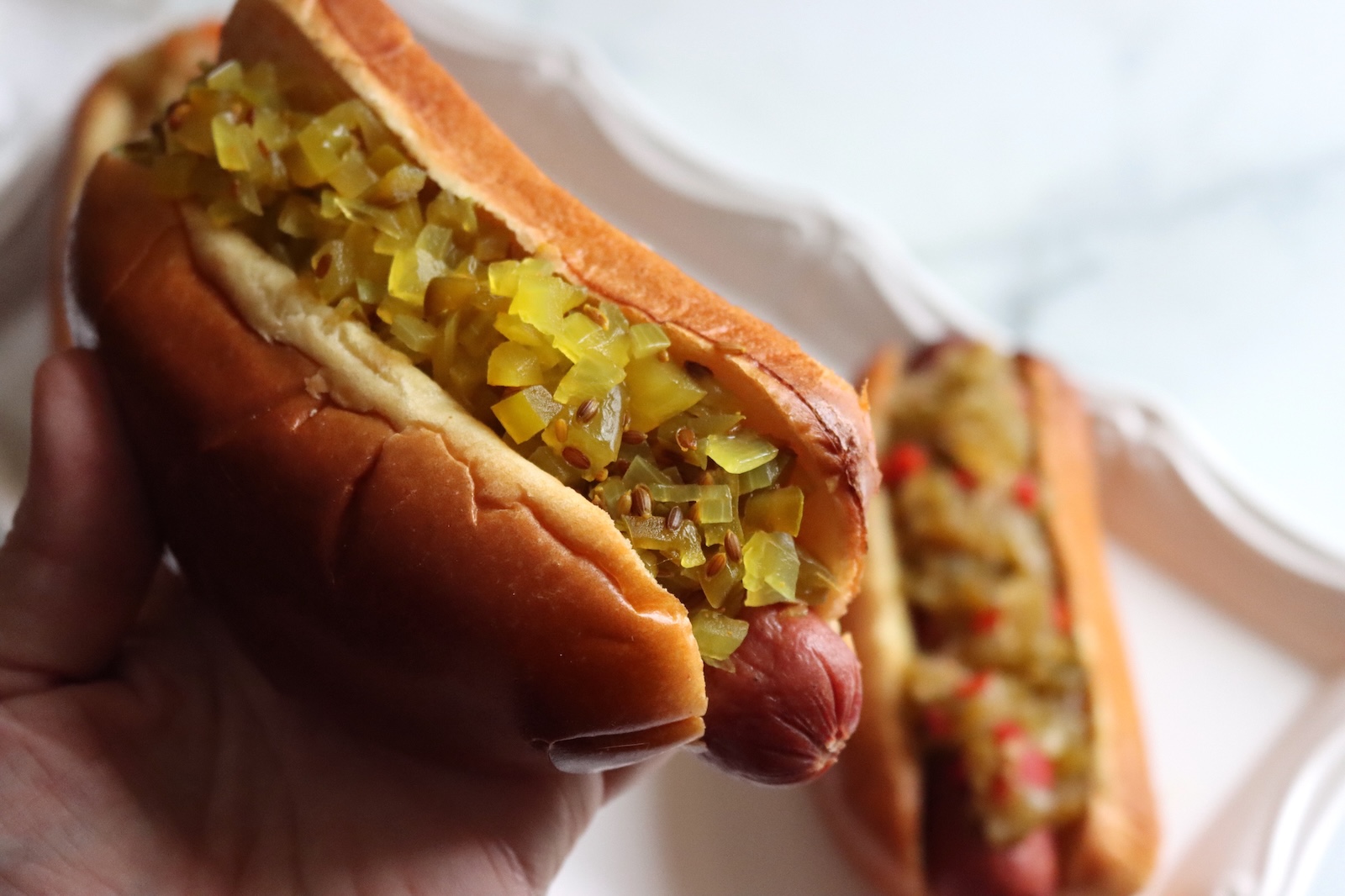 Hand holding a hot dog topped with dilled green tomato relish over a white platter