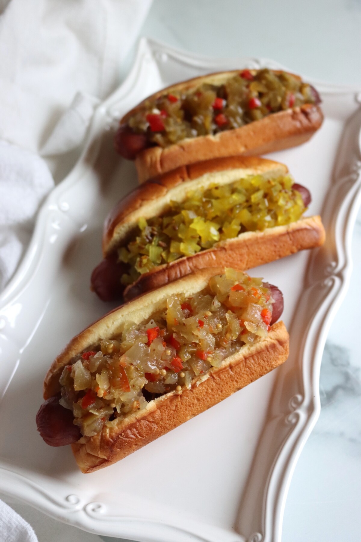 Hot dogs on a white platter topped with green tomato relish, the classic relish in front