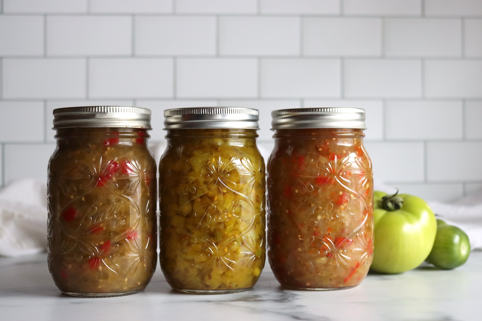Three pint jars of green tomato relish lined up next to whole green tomatoes