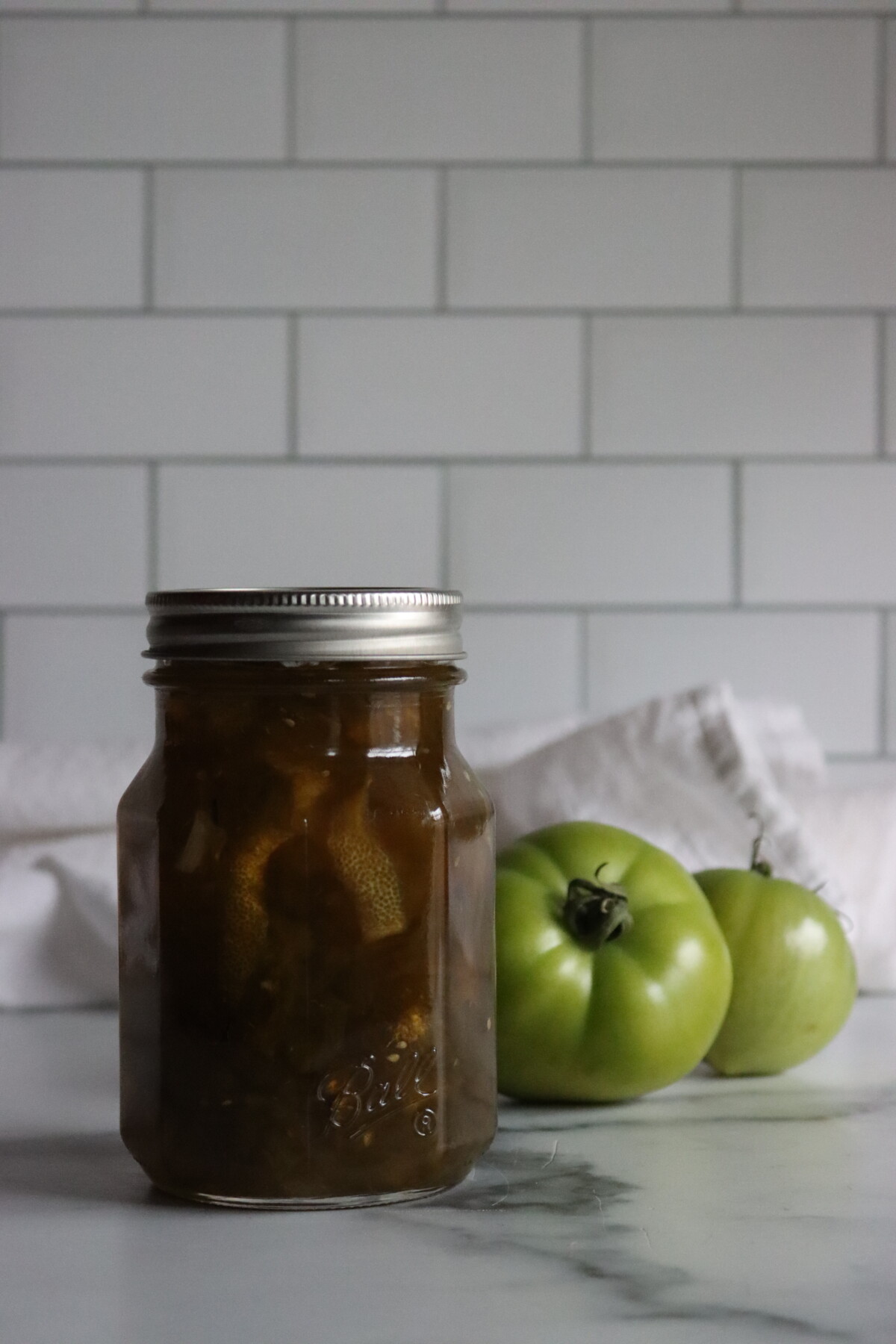 Sealed pint jar of green tomato preserves with green tomatoes behind it