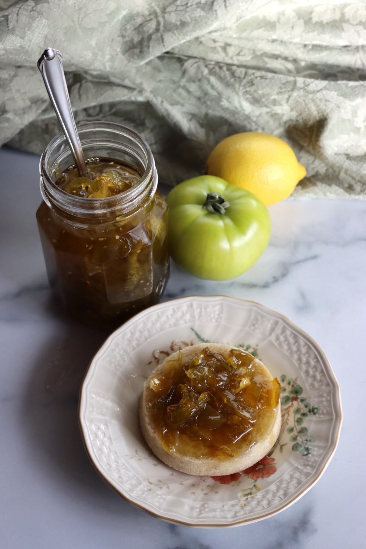 Open jar of Italian green tomato preserves with a spoon, and preserves spread on an English muffin