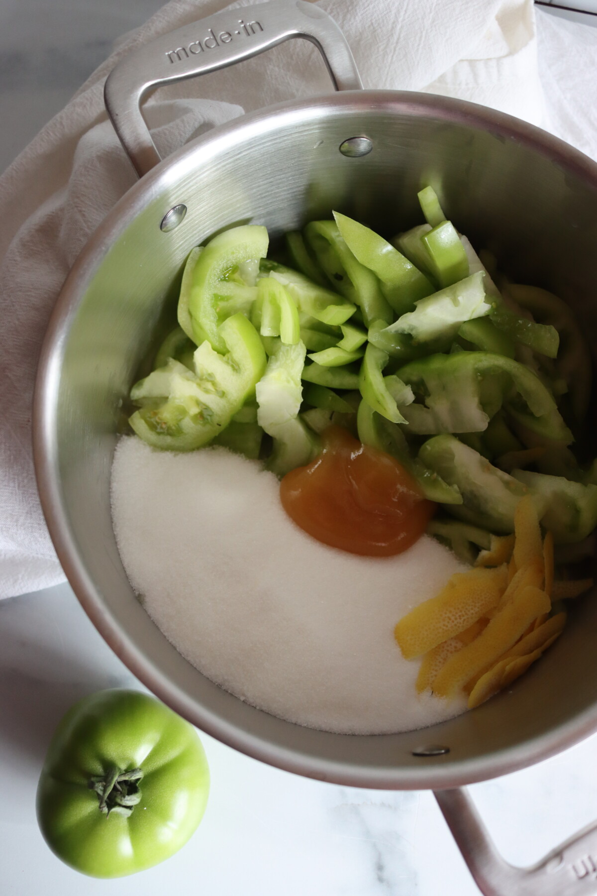 Sliced green tomatoes, sugar, honey, and strips of lemon peel in a stainless steel pot