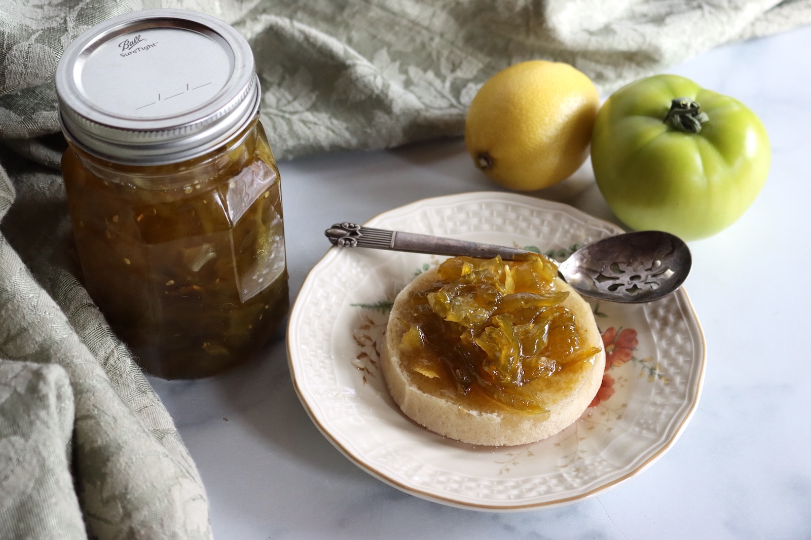 Jar of green tomato preserves with a spoonful on an English muffin, a lemon, and a green tomato