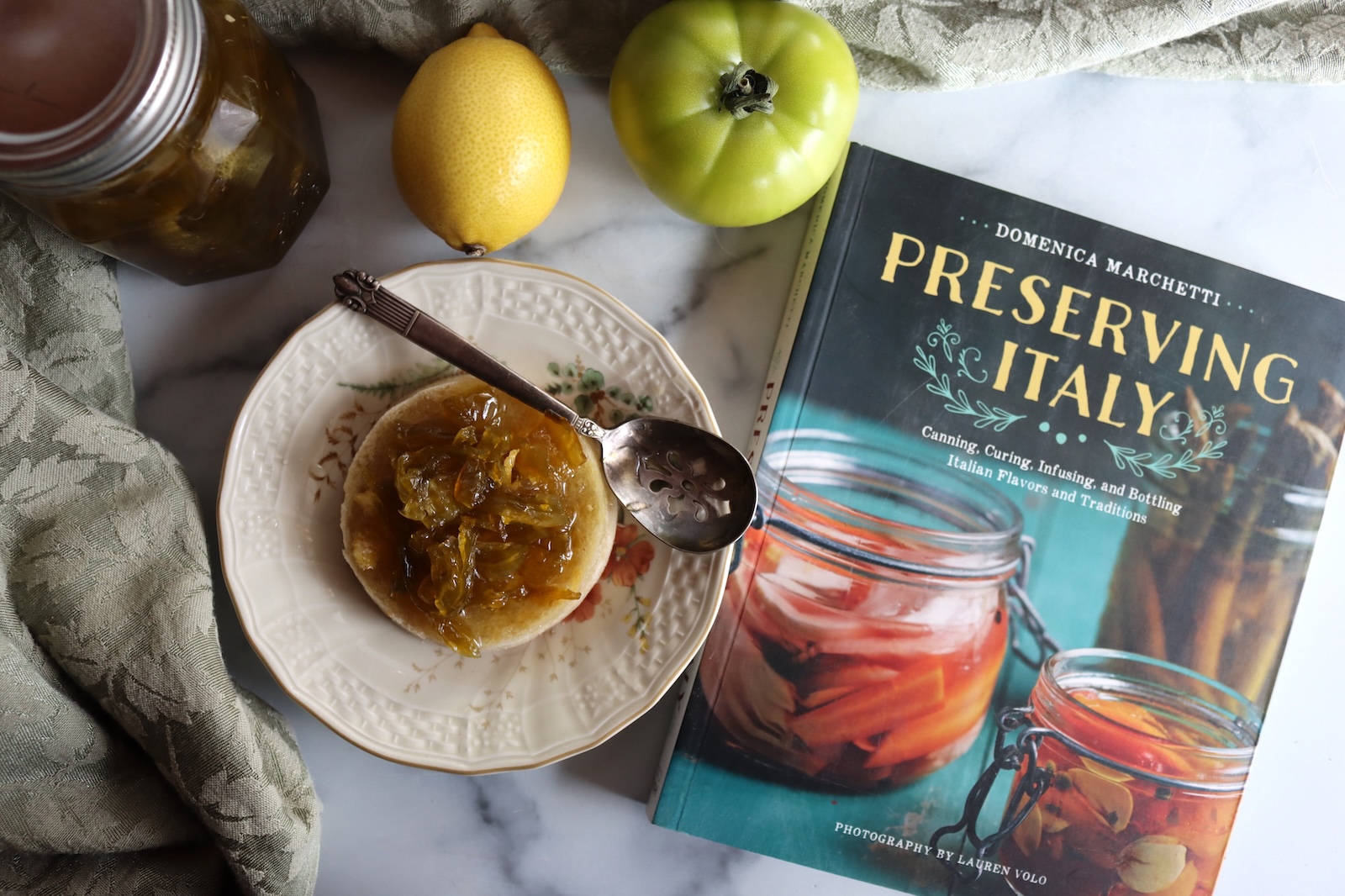 Green tomato preserves on an English muffin with a lemon, a green tomato, and the book Preserving Italy