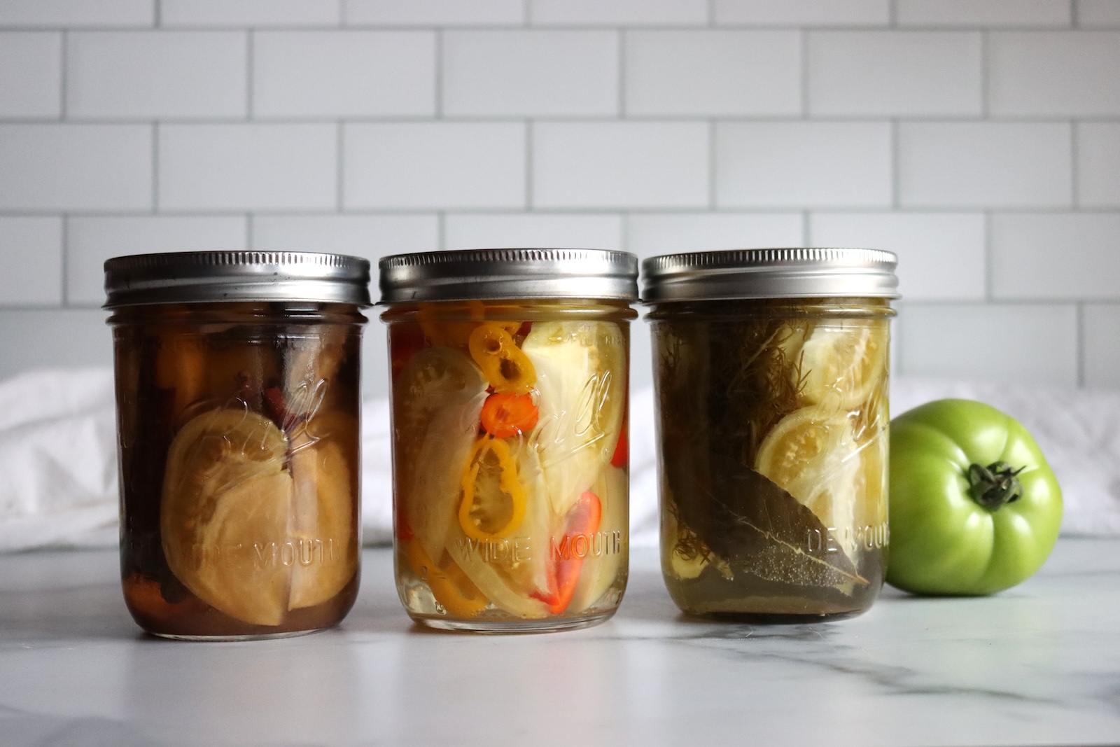 Three jars of sweet, spicy, and dill pickled green tomatoes beside a whole green tomato