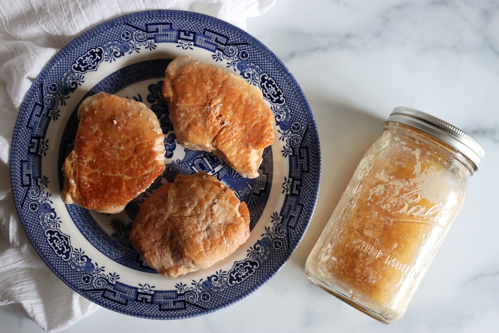 Three canned pork chops out of the jar on a blue and white plate, next to the jar of jellied juices