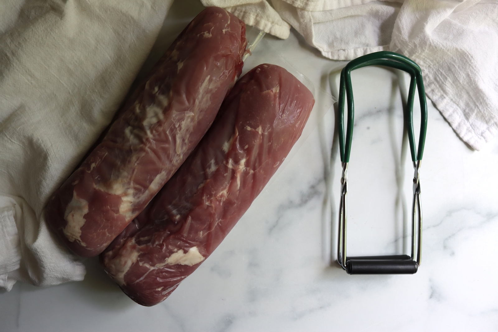 Two whole raw pork tenderloins on a marble counter next to a jar lifter