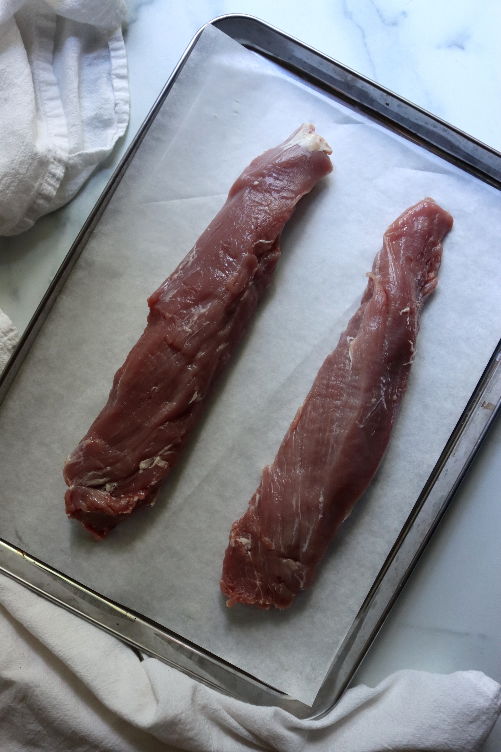 Two trimmed pork tenderloins on a parchment-lined baking sheet, ready to roast for a hot pack